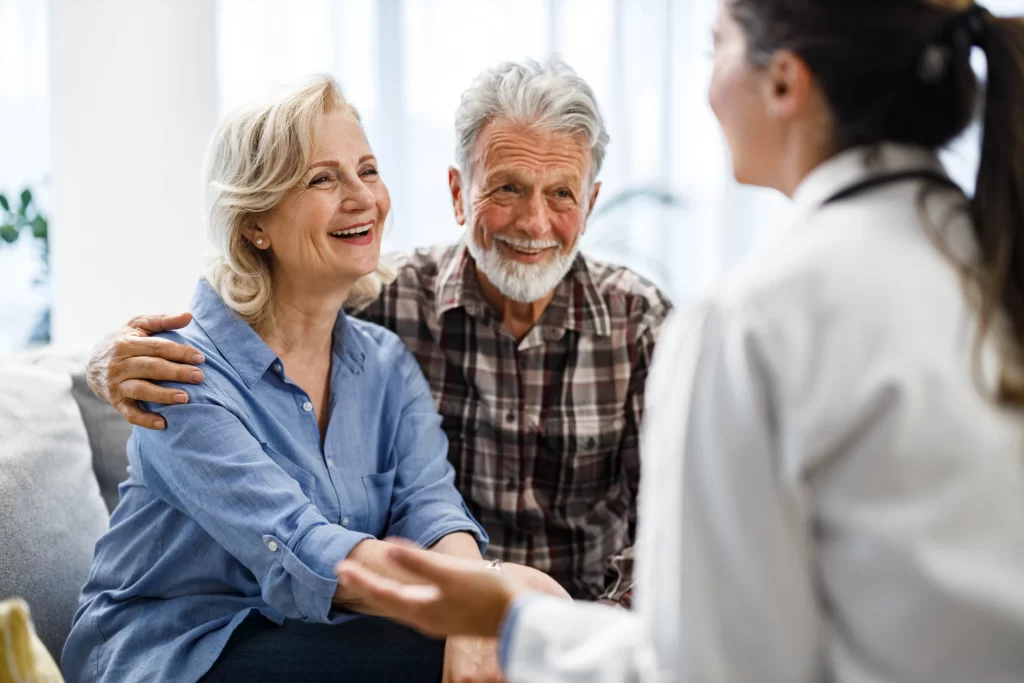 Smiling elderly couple sitting together on a sofa, speaking with a healthcare professional during a medical consultation.