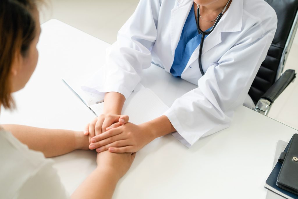 Doctor wearing a white coat gently holding a patient’s hands across a desk during a medical consultation.